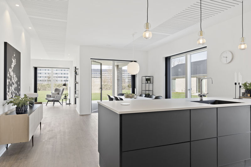 Moderne køkken-alrum fra eurodan-huse med mørke køkkenfronter, stor hvid bordplade, sort vask, flere små pendler over køkkenøen, akustikfelter i loftet, spiseplads ved vinduet og åbent kig til loungeområde – inspiration til stilrent nybyggeri og husdrømme med åbne rum.