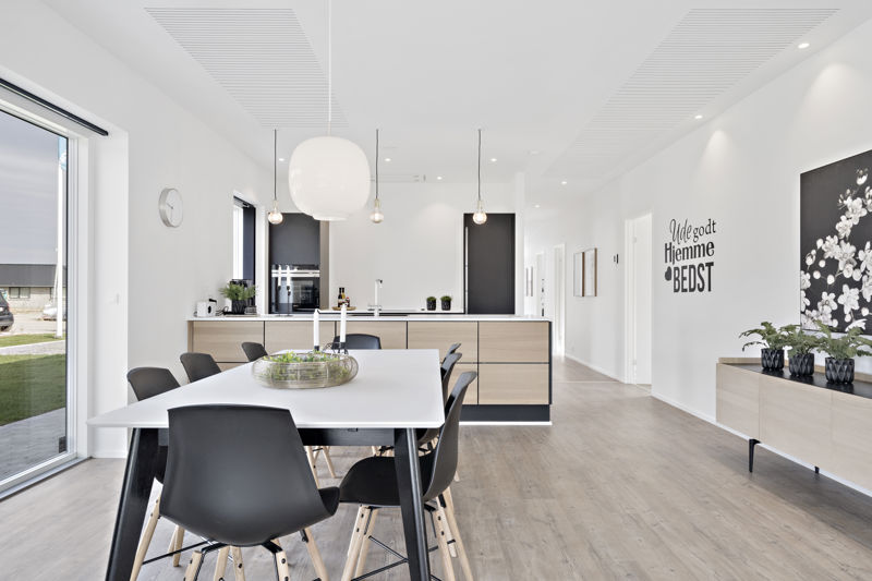 Lyst køkken-alrum fra eurodan-huse med hvidt spisebord, sorte stole, stor rund pendel, mindre pærer over køkkenet, køkkenø med træfronter, lang skænk med planter og vægtekst i gangen – inspiration til nybyggeri, husbyggeri og personlige husdrømme.