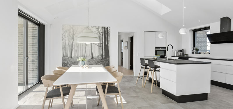 Køkken-alrum fra eurodan-huse med langt hvidt spisebord, lyse træstole, stor hvid pendel, vægbillede med skovmotiv, hvid køkkenø med sort bordplade, barstole og ovenlys i skråt loft – inspiration til nybyggeri, husbyggeri og lyse husdrømme.