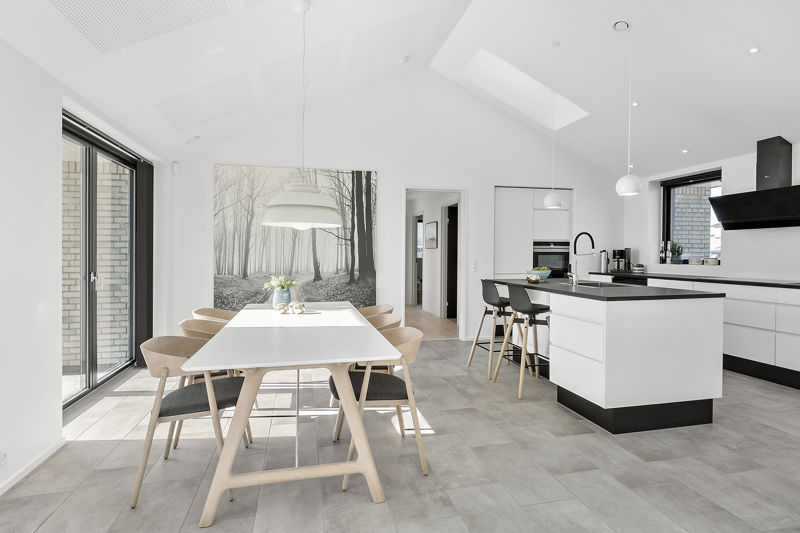 Køkken-alrum fra eurodan-huse med langt hvidt spisebord, lyse træstole, stor hvid pendel, vægbillede med skovmotiv, hvid køkkenø med sort bordplade, barstole og ovenlys i skråt loft – inspiration til nybyggeri, husbyggeri og lyse husdrømme.