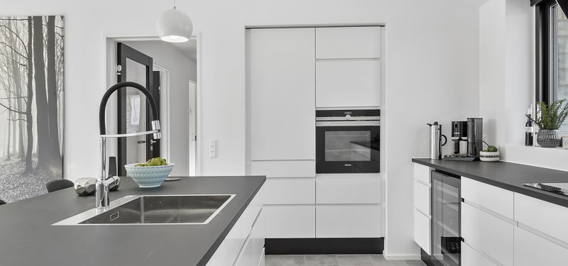 Detalje fra hvidt køkken fra eurodan-huse med sort bordplade, stor firkantet vask, markant sort armatur, integreret ovn i højskab, vinkøleskab under bordpladen og kaffestation ved vinduet – inspiration til funktionelt nybyggeri, husbyggeri og moderne køkkendrømme.