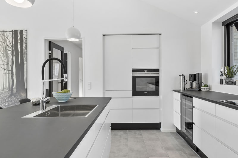 Detalje fra hvidt køkken fra eurodan-huse med sort bordplade, stor firkantet vask, markant sort armatur, integreret ovn i højskab, vinkøleskab under bordpladen og kaffestation ved vinduet – inspiration til funktionelt nybyggeri, husbyggeri og moderne køkkendrømme.