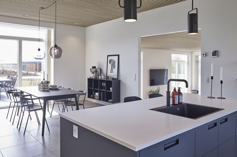 Køkken-alrum fra eurodan-huse med mørk køkkenø, hvid bordplade, sort vask og armatur, spiseplads med sort bord og stole, røgfarvede pendler, trælamelloft og åben forbindelse til stueområde – inspiration til moderne nybyggeri, husbyggeri og åbne husdrømme.
