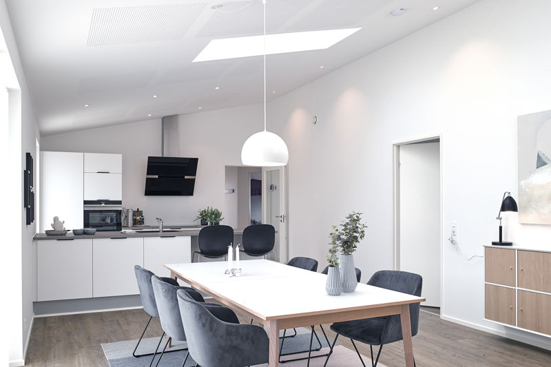 Lyst køkken-alrum fra eurodan-huse med hvidt spisebord, mørke polstrede stole, stor hvid pendel over spisepladsen, skråt loft med ovenlys, hvidt køkken med integreret ovn, sort emhætte og trælook gulv – inspiration til nybyggeri, husbyggeri og moderne husdrømme.