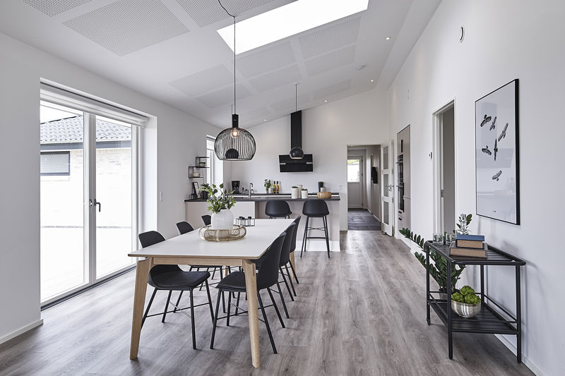 Åbent køkken-alrum fra eurodan-huse med hvidt spisebord, sorte stole, stor sort pendel, lange vinduespartier, barstole ved køkkenøen, sort emhætte, lys trægulvsoverflade og smalt konsolbord med planter på væggen – inspiration til nybyggeri, husbyggeri og åbne husdrømme.