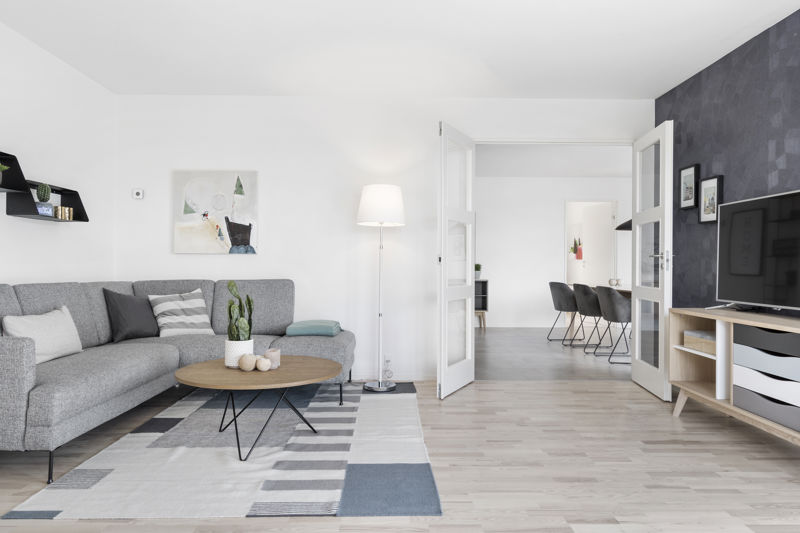 Stue fra eurodan-huse med grå sofa, rundt træsofabord, grafisk gulvtæppe, høj gulvlampe, tv-møbel foran mørk væg, åbne dobbeltdøre mod spiseplads og kig til køkken-alrum – inspiration til moderne nybyggeri, husbyggeri og funktionelle husdrømme.