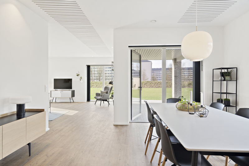 Åbent køkken-alrum og stue fra eurodan-huse med langt hvidt spisebord, sorte spisebordsstole, rund hvid pendel, sort reol, lyse trægulve, store terrassedøre og kig til tv-område i stuen – inspiration til nybyggeri, husbyggeri og åbne opholdsrum.