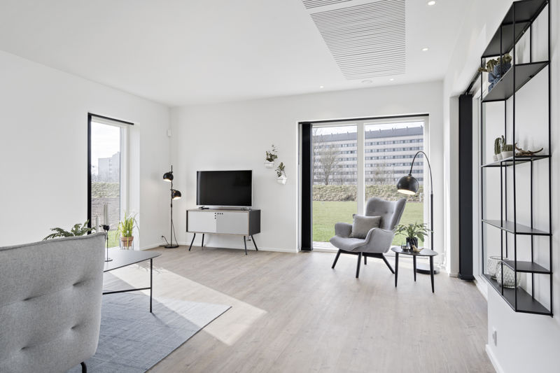Lys stue fra eurodan-huse med grå loungestol, tv på hvidt møbel, sort reol på væggen, buet gulvlampe, stort vinduesparti mod terrasse og akustikfelt i loftet – inspiration til nybyggeri, husbyggeri og moderne stueindretning.