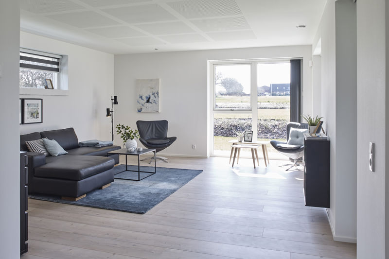 Stue fra eurodan-huse med mørk chaiselongsofa, blågråt gulvtæppe, lille glasbord, sort lænestol, lys loungestol ved vinduet, sort væghængt skænk og store vinduer med dagslys – konkret inspiration til moderne nybyggeri, husbyggeri og funktionelle husdrømme.