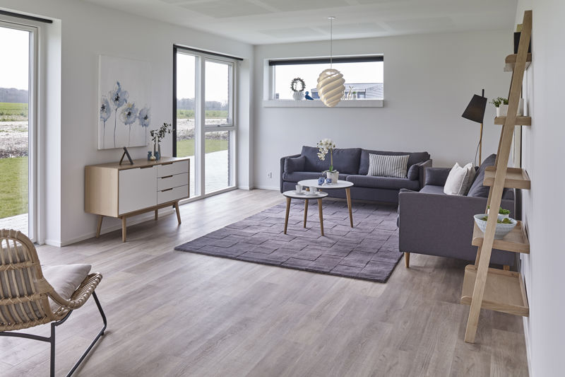 Lys stue fra eurodan-huse med mørkegrå sofa og chaiselong, rundt sofabord, lilla gulvtæppe, træstige-reol, lav skænk, kunstværk med blomster, flettet loungestol og store vinduer mod terrasse – inspiration til nybyggeri og husdrømme.