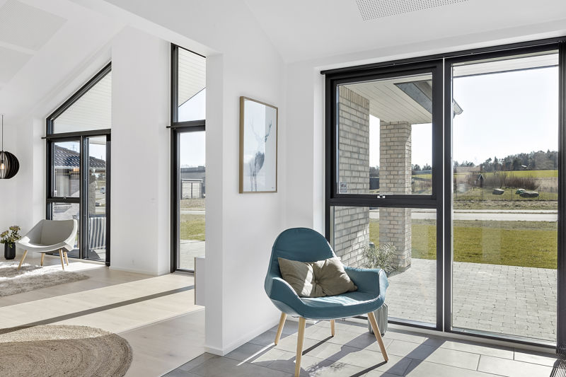 Stuehjørne fra eurodan-huse med blå loungestol, puder, sorte vinduesrammer, store gulv-til-loft vinduer, mørke klinker ved vinduespartiet og kig til lyst opholdsrum med høje gavlvinduer – inspiration til nybyggeri, husbyggeri og husdrømme med udsigt.