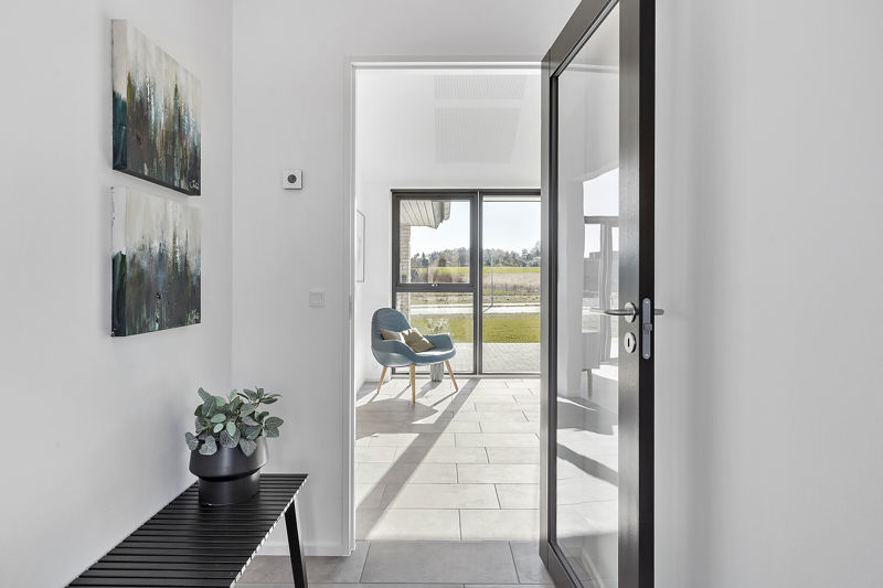 Kig fra entré mod stue fra eurodan-huse med sort bænk, grøn plante, to abstrakte billeder, høj glasdør med sort ramme, grå klinkegulv og blå loungestol ved stort vindue – inspiration til nybyggeri, husbyggeri og gennemtænkte husdrømme.