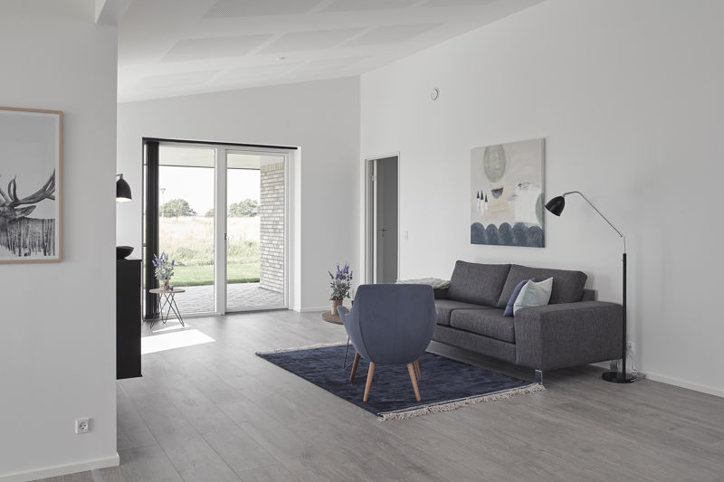Lys stue fra eurodan-huse med grå sofa, blå loungestol, mørkeblåt gulvtæppe, sort gulvlampe, stort vægmaleri, lille sidebord ved terrassedør og store vinduer mod overdækket terrasse – inspiration til nybyggeri, husbyggeri og hyggelige husdrømme.