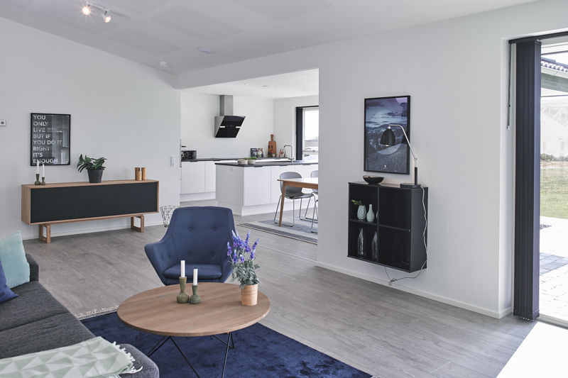 Stue fra eurodan-huse med blå loungestol, rund træ-sofabord, mørkeblåt tæppe, sort væghængt reol, sort skænk, sideboard i træ og åben forbindelse til hvidt køkken-alrum – inspiration til nybyggeri, husbyggeri og funktionelle opholdsrum.