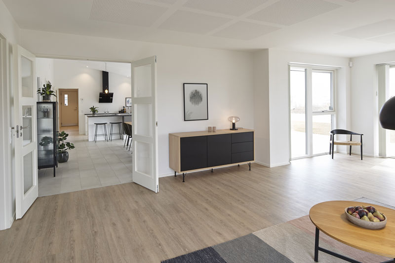 Åben stue fra eurodan-huse med lys trælook gulv, lav skænk med sorte fronter, dobbeltdøre mod køkken-alrum, sort vitrineskab med planter, rundt sofabord i forgrunden og stort vindue ved læsehjørne – inspiration til nybyggeri og husbyggeri.