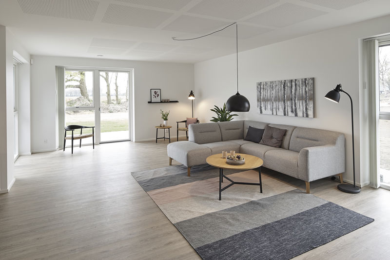 Stue fra eurodan-huse med lys grå hjørnesofa, rundt sofabord i træ, grafisk gulvtæppe, sort pendel, sort gulvlampe, vægkunst over sofaen og store vinduer på flere sider – inspiration til nybyggeri, husbyggeri og lyse opholdsrum.