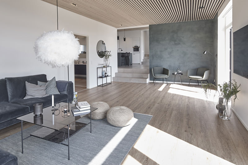 Stue fra eurodan-huse med mørk sofa, glas-sofabord, store strikkede puffer, gråt gulvtæppe, fluffy hvid pendel, akustikloft i trælameller, grå væg og hævet køkken i baggrunden – inspiration til nybyggeri, husbyggeri og moderne husdrømme.