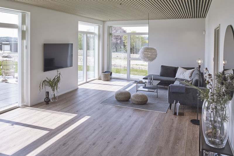 Lys stue fra eurodan-huse med grå sofa, firkantet sofabord, to runde puffer, væghængt tv, stort glasparti mod terrasse, åben dør til uderum og lamelloft med trædetaljer – konkret inspiration til nybyggeri, husbyggeri og lyse husdrømme.