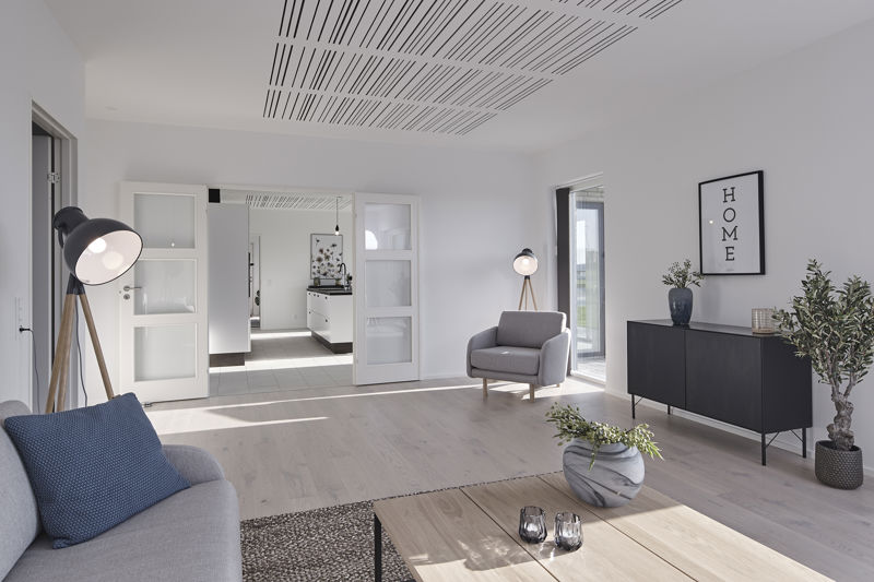 Rummelig stue fra eurodan-huse med lyse trægulve, grå sofa, stor sort gulvlampe, grå loungestol, sort skænk, plantekrukker, dobbelt døråbning mod entré og akustikloft med lange sorte spor – inspiration til moderne nybyggeri og funktionelle husdrømme.