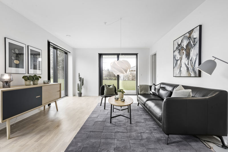 Stue fra eurodan-huse med sort lædersofa, rund træbordplade på sort metalstel, gråt tæppe, mørk loungestol, stor hvid pendel, lav skænk i træ og sort front samt store vinduer med kig til have – inspiration til stilrent nybyggeri, husbyggeri og moderne husdrømme.