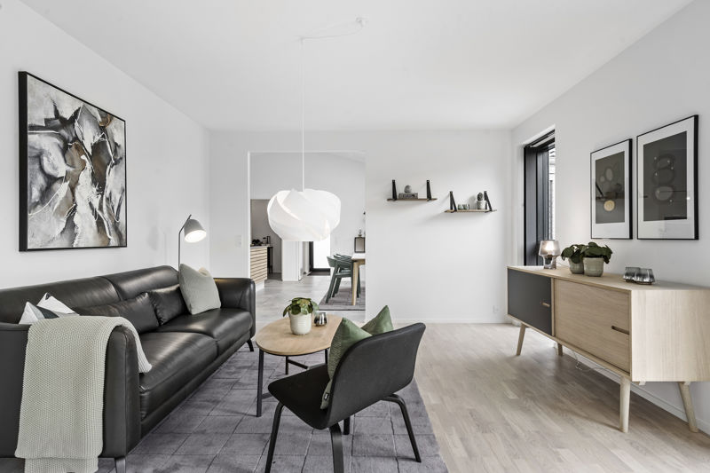 Stue fra eurodan-huse med sort lædersofa, mørk loungestol med grøn pude, rundt sofabord i træ, gråt gulvtæppe, stor hvid pendel, lav skænk i træ med sorte fronter og væghylder i baggrunden – konkret inspiration til nybyggeri, husbyggeri og personlige husdrømme.