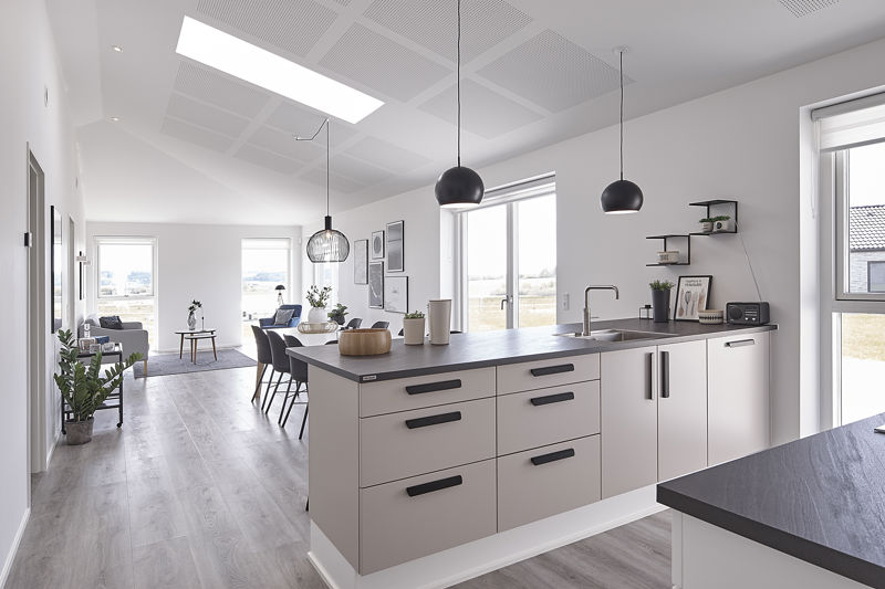 Åbent køkken-alrum og stue fra eurodan-huse med lyst køkken, sort bordplade, sorte greb, spiseplads, opholdsstue i baggrunden, store vinduer, ovenlys og sorte pendler over køkkenøen – inspiration til nybyggeri, husbyggeri og åbne opholdsrum.