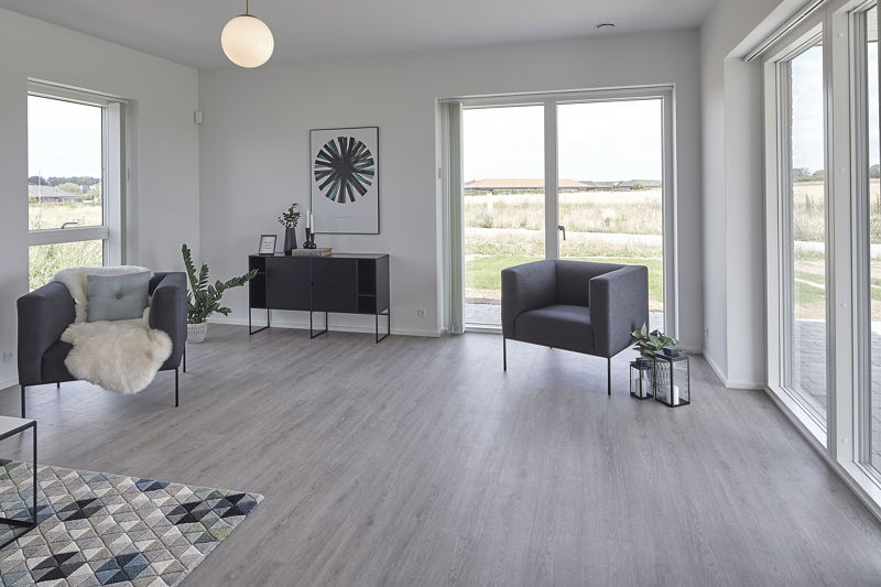 Lys stue fra eurodan-huse med to mørke loungestole, sort skænk, rund hvid pendel, geometrisk gulvtæppe, plante ved væggen og store vinduespartier på to sider med direkte udgang til terrasse – inspiration til nybyggeri, husbyggeri og lyse husdrømme.