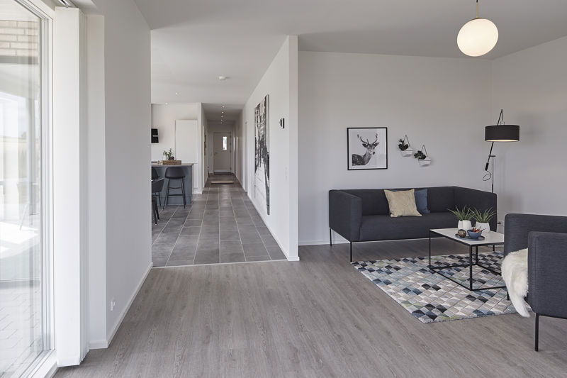 Stue fra eurodan-huse med mørk sofa, mørk loungestol med lammeskind, sort sofabord, mønstret gulvtæppe, væglampe, rund hvid loftlampe og åben forbindelse til gang og køkken-alrum – inspiration til funktionelt nybyggeri, husbyggeri og sammenhængende husdrømme.