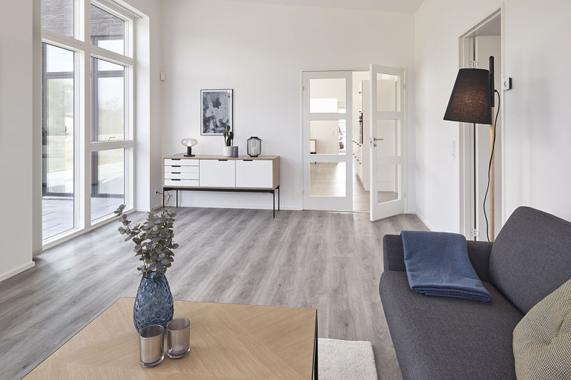 Stue fra eurodan-huse med mørkegrå sofa, stort lyst sofabord i forgrunden, blå vase, høj sort gulvlampe, hvid skænk med træramme, store vinduer og dobbeltdør mod resten af boligen – inspiration til nybyggeri, husbyggeri og moderne opholdsrum.