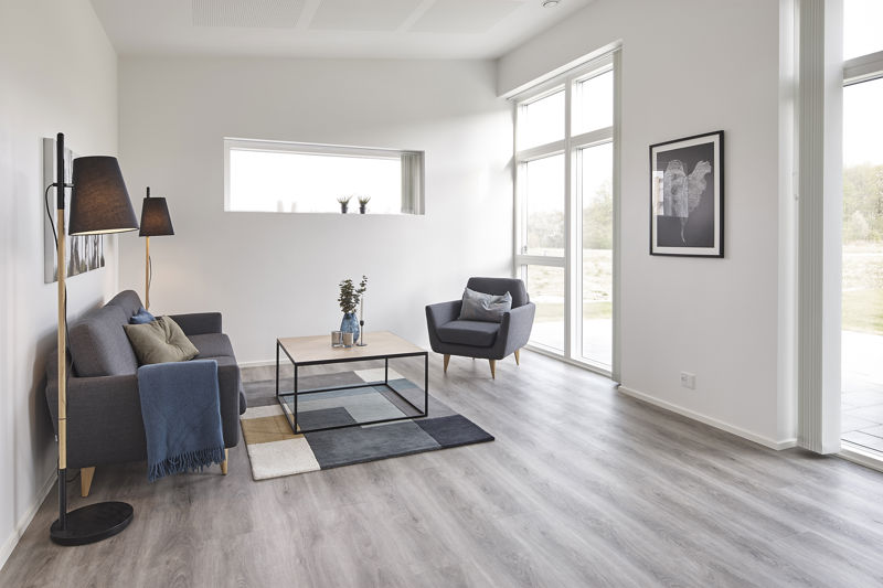Lys stue fra eurodan-huse med mørkegrå sofa, matchende lænestol, firkantet sofabord, patchwork-gulvtæppe, høj gulvlampe, stort hjørnevindue og smalt vandret vindue med små planter i karmen – inspiration til nybyggeri, husbyggeri og lyse husdrømme.