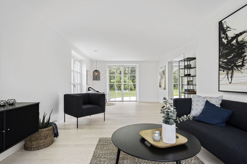 Stue fra eurodan-huse med mørk sofa, sort rundt sofabord, stort naturtæppe, sort skænk, fletkurv med plante, mørk lænestol, reol som rumdeler og sprossede terrassedøre med kig til have – inspiration til nybyggeri, husbyggeri og stue med karakter.