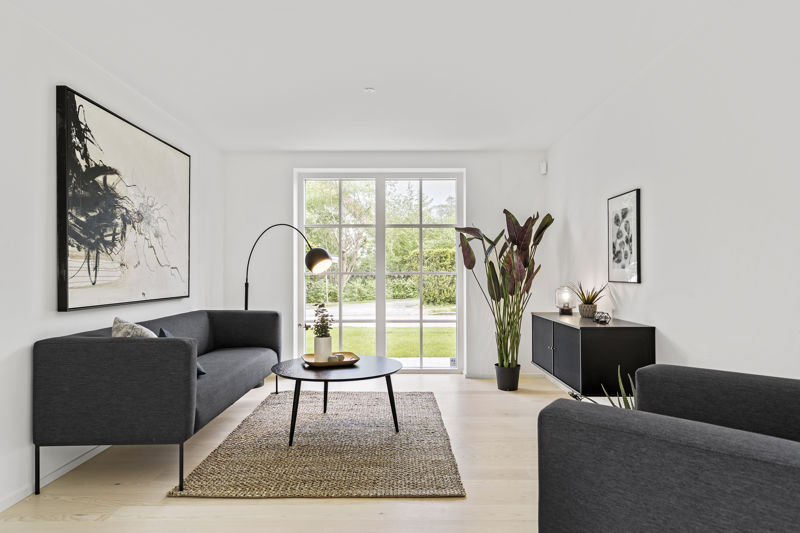 Symmetrisk stue fra eurodan-huse med to mørke sofaer overfor hinanden, sort rundt sofabord, buet gulvlampe, stort naturtæppe, sort skænk, høj plante og sprossede terrassedøre mod haven – inspiration til klassisk nybyggeri, husbyggeri og rolige husdrømme.