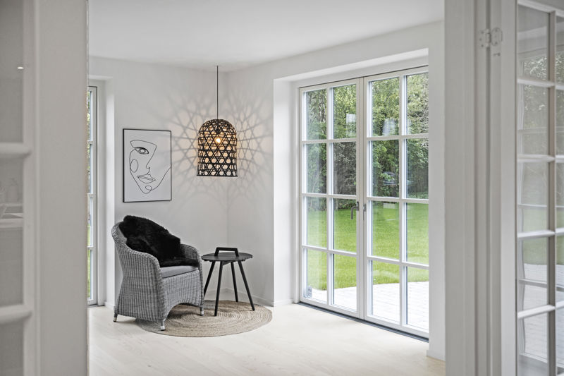 Hyggeligt stuehjørne fra eurodan-huse med flettet loungestol, sort lammeskind, rundt jute-tæppe, lille sort sidebord, dekorativ sort pendel med lysmønster på væggen og store sprossede terrassedøre mod haven – inspiration til nybyggeri, husbyggeri og personlige husdrømme.