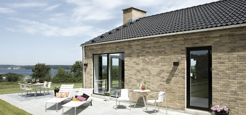 Moderne murstenshus med sort saddeltag, store vinduespartier og solrig fliseterrasse med udsigt over vandet – inspiration til udeliv ved et nyt eurodan-hus.