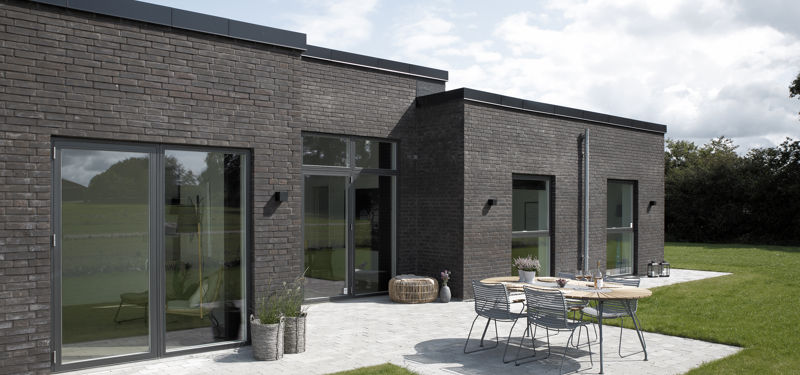 Moderne H-hus med mørk murstensfacade, fladt tag, store vinduespartier og flisebelagt terrasse med havemøbler ud mod en grøn have – stilren inspiration til nybyg og husdrømme.