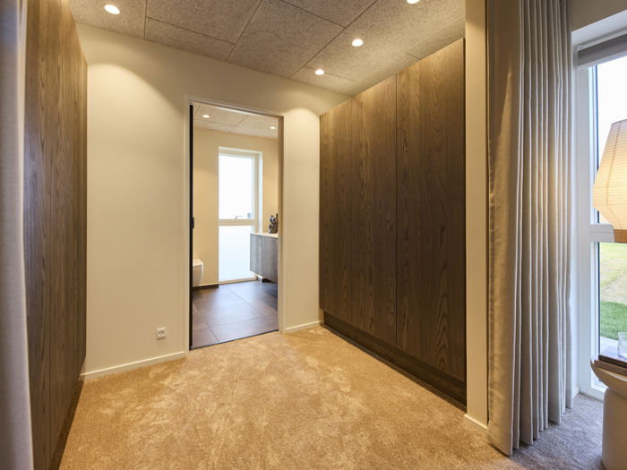 Walk-in-closet i moderne vinkelhus med mørke garderobeskabe, blødt gulvtæppe og direkte adgang til eget badeværelse – inspiration til drømmehuset.