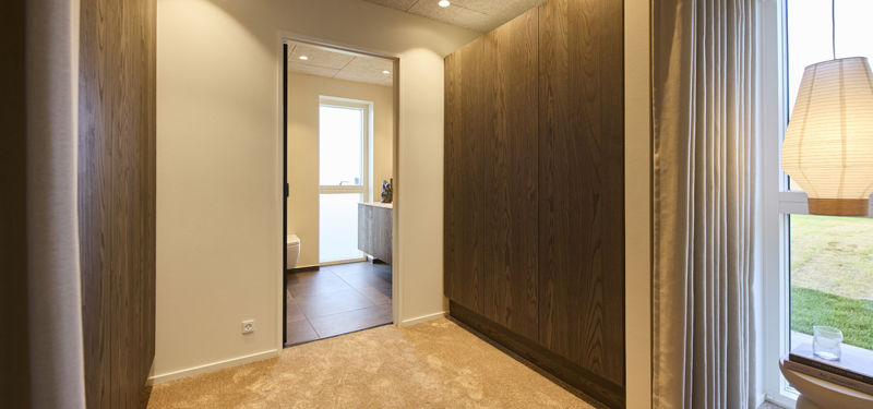 Walk-in-closet i moderne vinkelhus med mørke garderobeskabe, blødt gulvtæppe og direkte adgang til eget badeværelse – inspiration til drømmehuset.