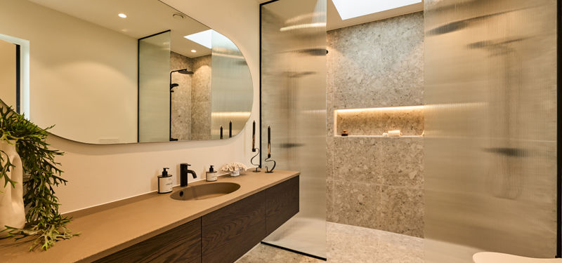 Badeværelse fra eurodan-huse med langt badmøbel i mørkt træ, beige bordplade, sort armatur, rundt spejl, ovenlys, glasvægge med struktur og bruseniche med terrazzo-look klinker – inspiration til moderne nybyggeri og badeværelsesdesign.