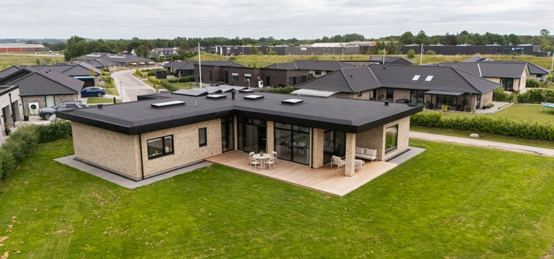 Moderne funkishus fra eurodan-huse med lys murstensfacade, sort fladt tag, store vinduespartier, græsplæne og træterrasse rundt om boligen – inspiration til nybyggeri, husbyggeri og arkitekttegnede husdrømme.