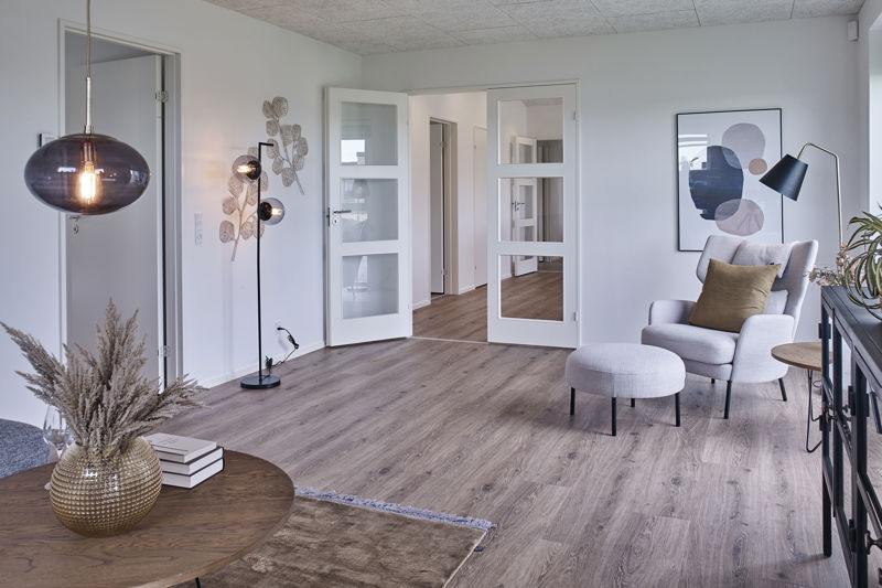 Stue fra eurodan-huse med lys lænestol og rund puf, sort vitrineskænk, rundt træbord, stor røgfarvet pendel, sort standerlampe med glaskupler, dobbeltdøre mod gang og trælook gulv – inspiration til nybyggeri, husbyggeri og funktionel stueindretning.