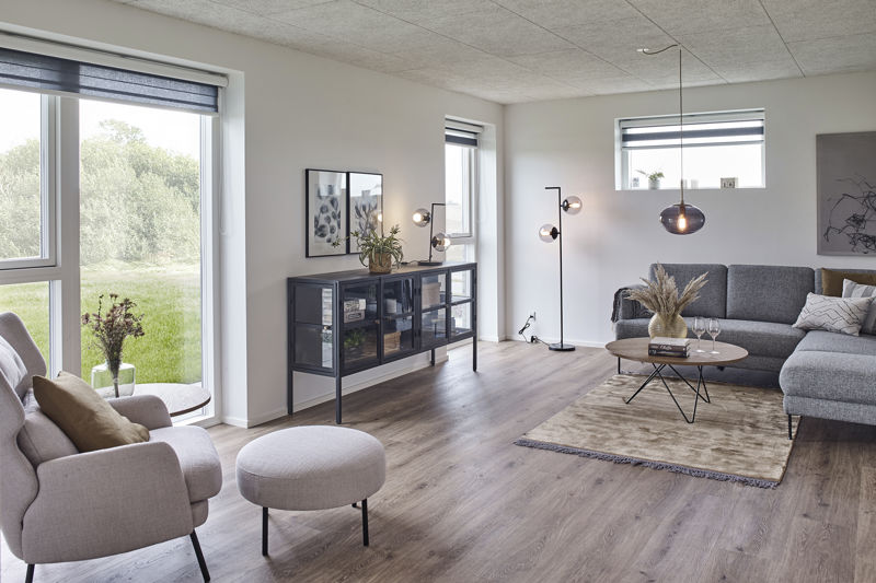 Stue fra eurodan-huse med grå hjørnesofa, rund puf, rundt sofabord, sort vitrineskænk, standerlampe, mørke rullegardiner og store vinduer med udsyn til grønt område – inspiration til nybyggeri, husbyggeri og komfortable husdrømme.