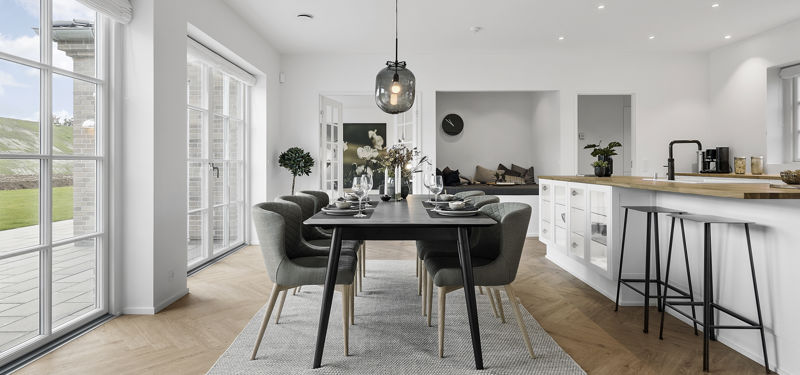 Lyst køkken-alrum i et moderne nybyggeri med spisebord, hvide køkkenelementer, køkkenø med barstole, store vinduespartier og sildebensgulv.