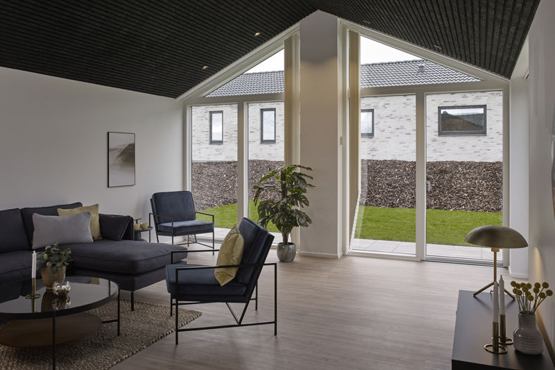 Rummelig stue fra eurodan-huse med sort akustikloft, mørk sofa, sorte loungestole, rundt sofabord, stor grøn plante, høje gavlvinduer og brede terrassedøre med kig til haven – inspiration til nybyggeri, husbyggeri og husdrømme med store vinduespartier.