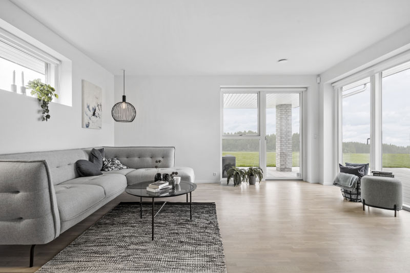 Lys stue fra eurodan-huse med lang grå sofa, rundt sort sofabord, groft gråt gulvtæppe, sort pendel, højt smalt vindue med plante, store terrassevinduer og læsehjørne ved vinduet – inspiration til nybyggeri, husbyggeri og rolige husdrømme.