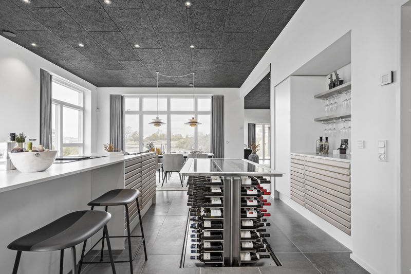 Køkken-alrum fra eurodan-huse med barstole ved køkkenøen, indbygget vinopbevaring i gulvet, åben barhylde med glas, trælamel-fronter, mørkt akustikloft, store vinduer og spiseplads for enden af rummet – inspiration til nybyggeri, husbyggeri og gennemtænkte køkkenløsninger.