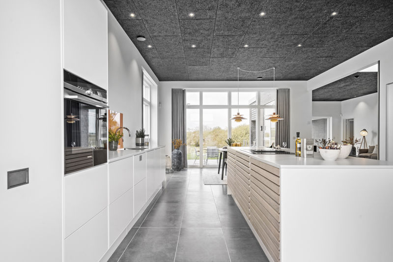 Langt køkken fra eurodan-huse med hvid skabsvæg, integreret ovn, mørkt akustikloft, grå klinkegulv, trælamel-køkkenø med hvid bordplade, stor vinduesfacade ved spisepladsen og indbygget barhylde i væggen – inspiration til funktionelt nybyggeri, husbyggeri og moderne køkken-alrum.