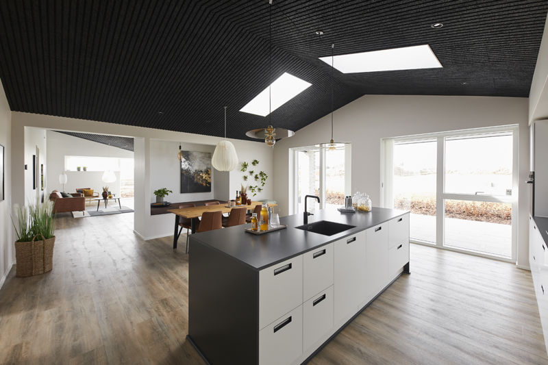 Åbent køkken-alrum fra eurodan-huse med stor køkkenø med mørk bordplade, sort vask og armatur, hvide skuffer, sort loft med ovenlys, spiseplads bag køkkenet og store terrassevinduer mod haven – inspiration til moderne nybyggeri, husbyggeri og køkken-alrum med dramatisk loftsudtryk.