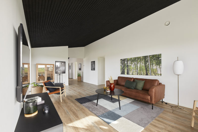 Stue fra eurodan-huse med cognacfarvet sofa, sort rundt sofabord, grafisk gulvtæppe, sort akustikloft, stor skovplakat, hvid gulvlampe, sort tv-møbel og kig mod spiseplads – inspiration til nybyggeri, husbyggeri og moderne husdrømme.