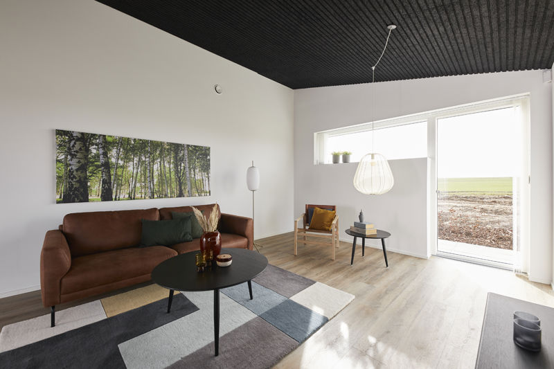 Stue fra eurodan-huse med cognacfarvet sofa, sort rundt sofabord, grafisk gulvtæppe, stor skovplakat, sort akustikloft, hvid pendel, loungestol med gul pude og stort vindue mod landskab – inspiration til nybyggeri, husbyggeri og varme husdrømme.