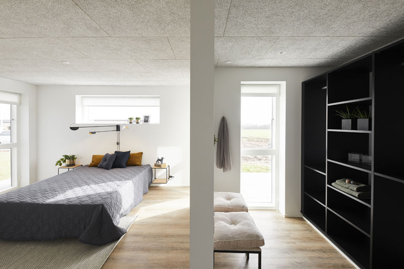 Soveværelse og walk-in fra eurodan-huse med seng, gråt sengetæppe, sengebord med plante, høj vinduesplacering, bænk ved skillevæg og sort åben garderobereol med hylder – inspiration til moderne opbevaring i nybygget hus og husdrømme.